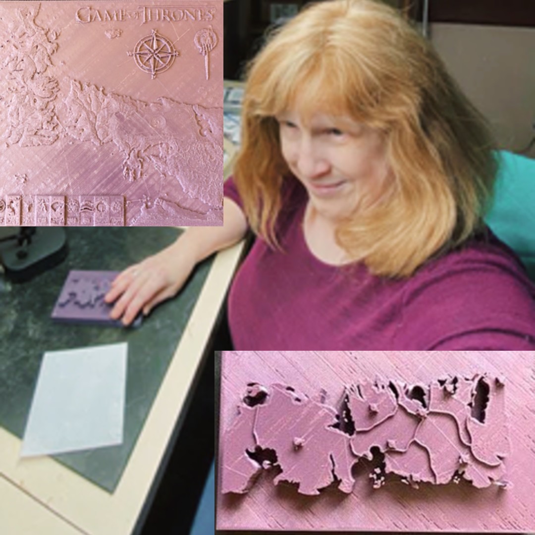 Jenine is sitting at a desk with her Game of Thrones models displayed in front of her.  She is feeling the purple model that shows all of the continents in the GOT series.  Jenine is smiling softly, wearing a plum purple shirt, and has strawberry blonde hair. 